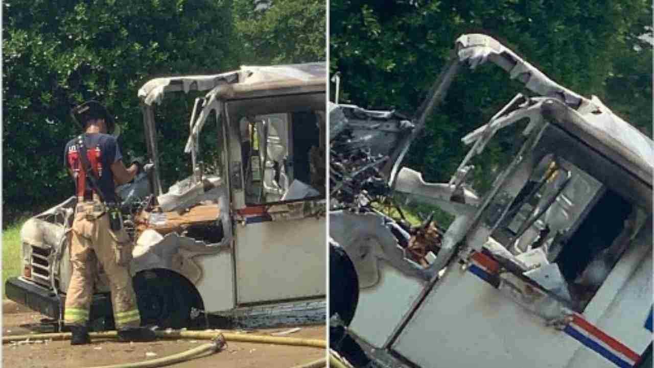 A firefighter examining a damaged postal delivery truck with significant fire damage to the front. The scene is in a suburban area with green trees in the background.