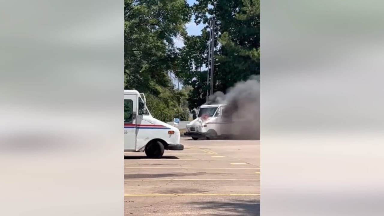 A postal truck emitting dark smoke in a parking lot with another postal truck visible in the foreground. Green trees are seen in the background under a clear sky.