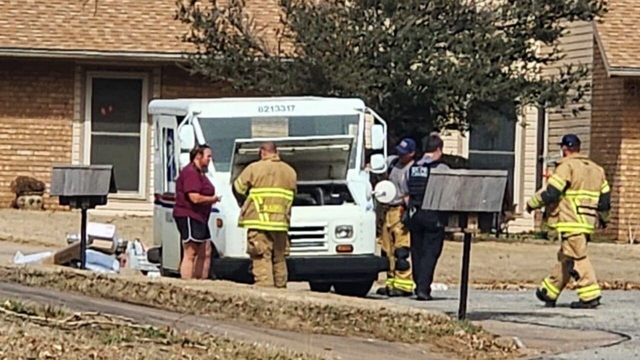 Emergency responders assisting a woman near a delivery truck outside a residence. Firefighters and police are present, with materials scattered on the ground.