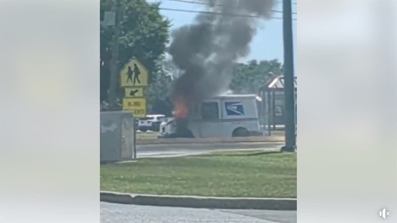 A postal service delivery vehicle emitting black smoke, with flames visible at the rear, parked near a street and pedestrian crossing sign. The scene is set in a suburban area with other vehicles in the background.
