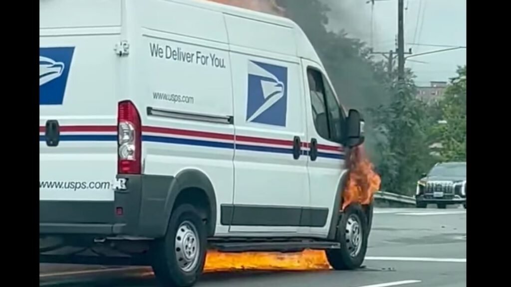 A USPS delivery van parked on a road with flames visible on the rear side, indicating a fire. Smoke is rising from the vehicle, and a nearby car is seen in the background.