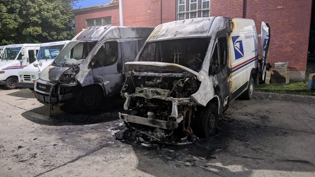 Two burnt delivery vans parked in a lot, one displaying a postal service logo, with visible damage to the front and sides from fire. The surrounding area shows a clear sky and a brick building in the background.