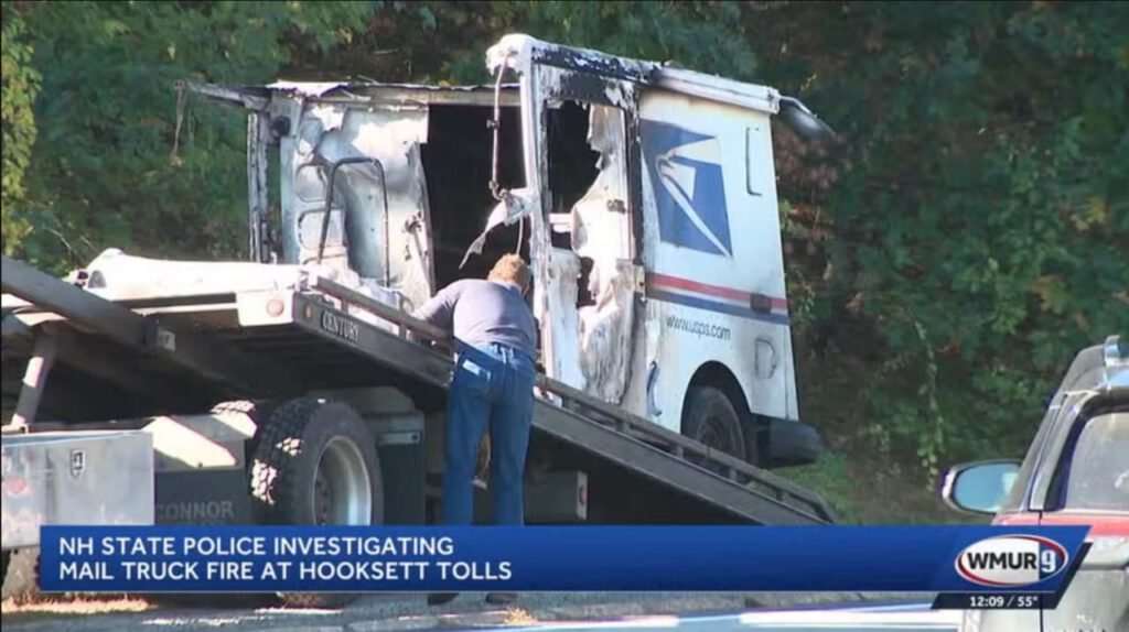 A damaged United States Postal Service truck being loaded onto a tow truck after a fire, with a state police officer investigating the scene. Dense greenery in the background.