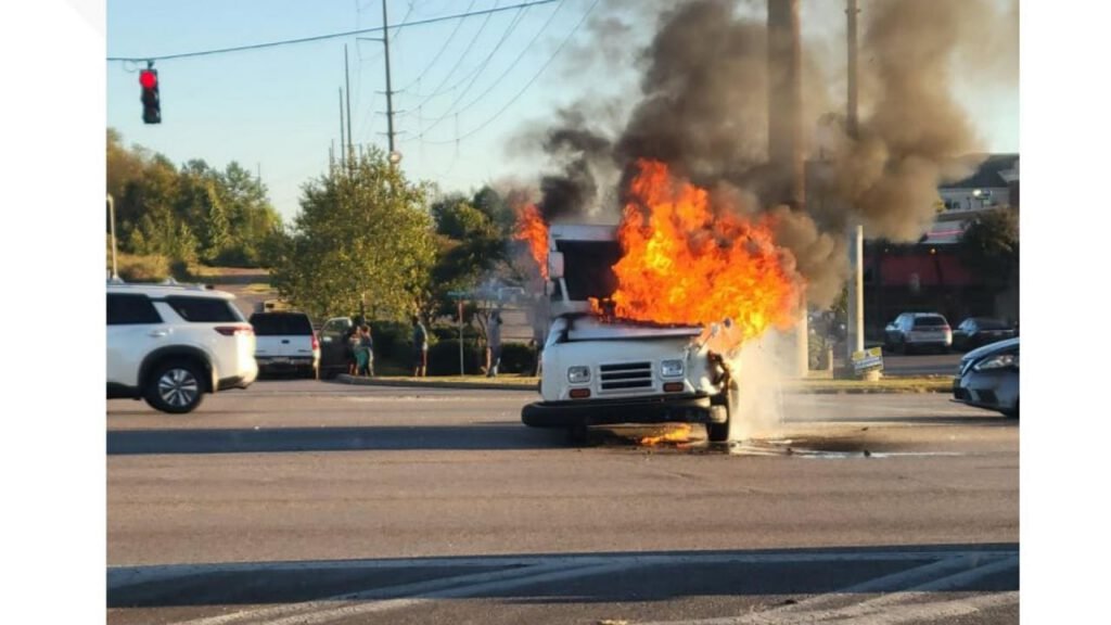 A vehicle on fire in the middle of an intersection, with flames and smoke rising from the front. Cars are stopped nearby, and traffic lights are visible in the background.