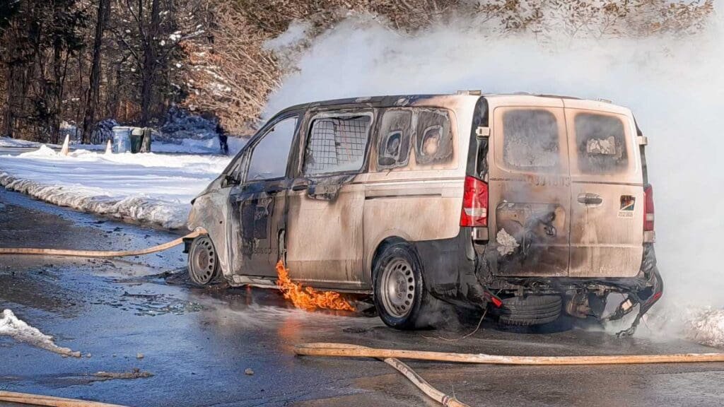 A burnt-out van with smoke rising from it on a snowy road, with fire hoses laid out nearby. The vehicle shows signs of fire damage with charred areas and exposed metal.