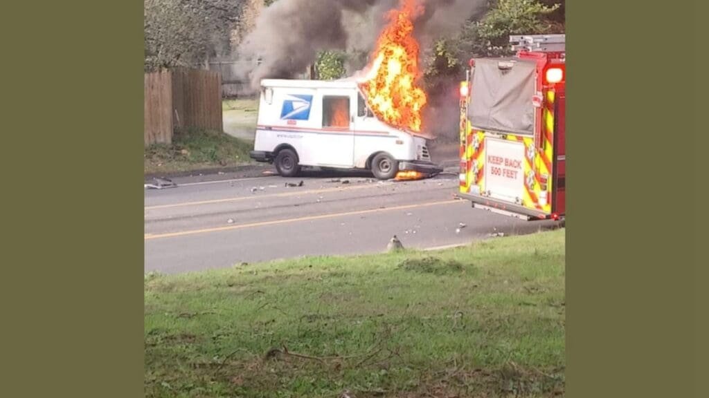 A postal service vehicle engulfed in flames on a street, with smoke billowing from the front. A fire truck with emergency lights is parked nearby, responding to the incident.