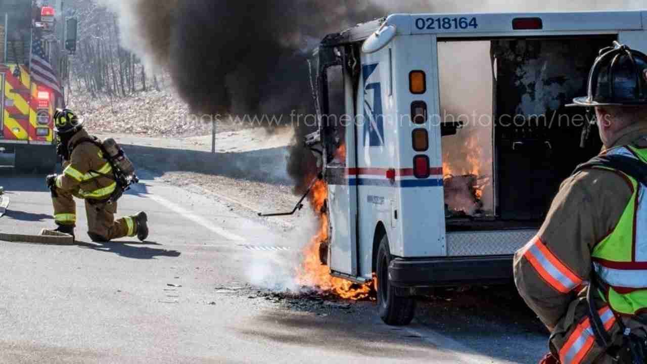 A fire truck parked beside a burning postal vehicle. Firefighters are responding, with one kneeling near the flames and smoke emanating from the vehicle's rear. The scene shows a road with dry surroundings.