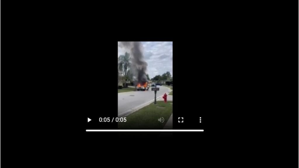 A vehicle on fire with thick smoke rising into the air, located in a residential area. A fire hydrant is visible nearby, and houses are in the background under a cloudy sky.