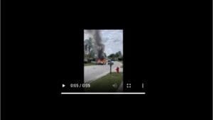 A vehicle on fire with thick smoke rising into the air, located in a residential area. A fire hydrant is visible nearby, and houses are in the background under a cloudy sky.