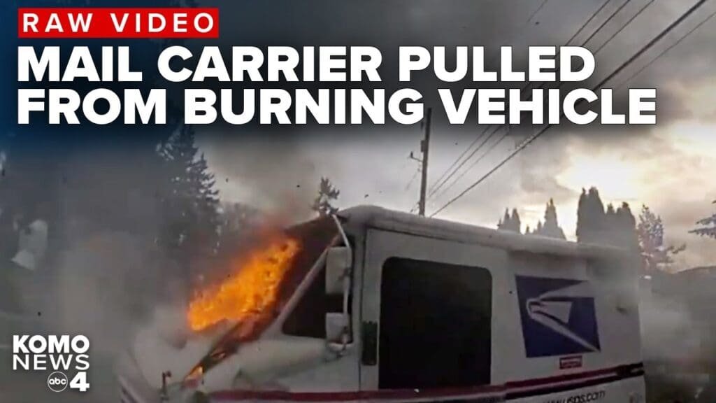 A mail delivery vehicle engulfed in flames with smoke rising in the background. The scene is set on a road with trees nearby and utility poles in the distance. Text overlay reads 'RAW VIDEO MAIL CARRIER PULLED FROM BURNING VEHICLE'.