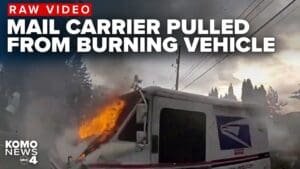 A mail delivery vehicle engulfed in flames with smoke rising in the background. The scene is set on a road with trees nearby and utility poles in the distance. Text overlay reads 