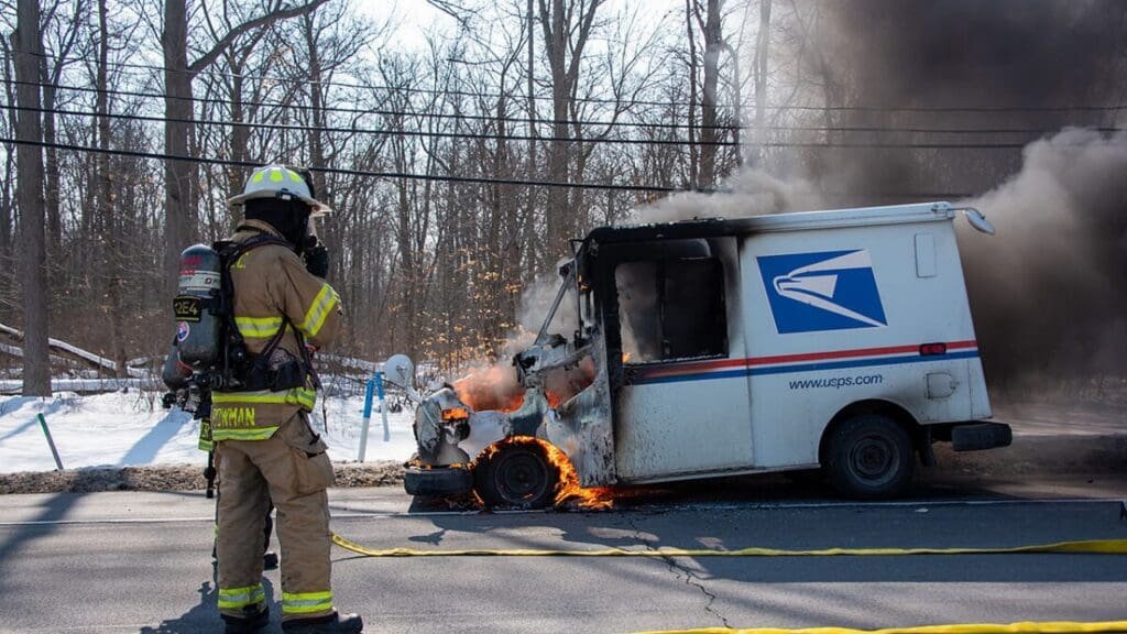 A firefighter monitoring a USPS delivery truck that is on fire on the side of a snow-covered road, with smoke billowing from the vehicle in a wooded area.