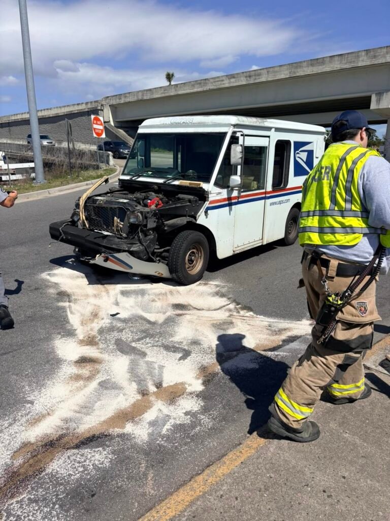 A damaged USPS delivery van on a road, with visible front-end damage. Emergency responders in safety gear are present, and a white powder is spread on the roadway, likely for cleanup. A highway overpass is visible in the background.