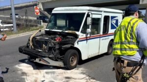 A damaged USPS delivery vehicle on the road with its hood open, showing the engine. A person in a safety vest is inspecting the vehicle. The background includes a stop sign and an overpass.