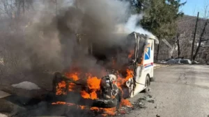 A postal delivery vehicle engulfed in flames, with thick black smoke rising from it on a street. Charred debris surrounds the vehicle, and trees are visible in the background.