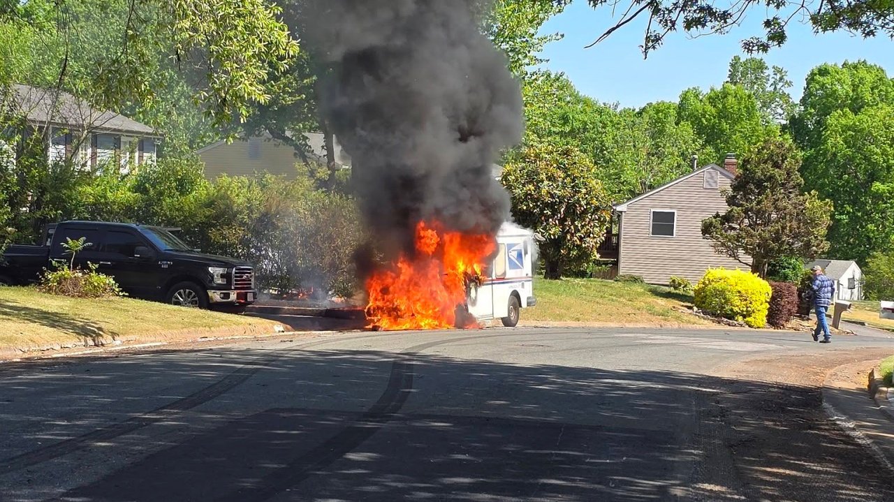 A burning vehicle with visible flames and dense black smoke rising in a residential area. Nearby houses are visible, along with a parked black truck. A person is seen walking away from the scene, while the road appears damaged from tire marks.