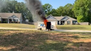 A postal delivery truck is on fire, emitting large clouds of black smoke. The scene is set outside residential houses with green trees in the background. The truck