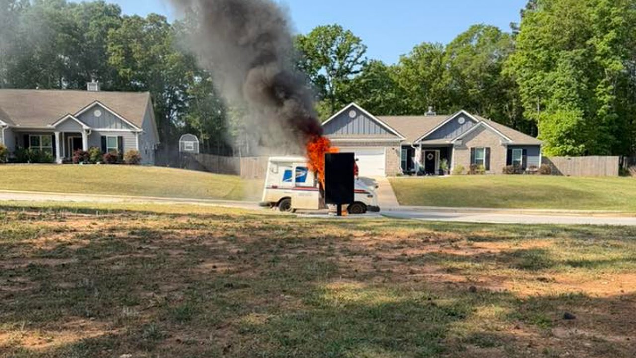 A postal delivery truck is on fire, emitting large clouds of black smoke. The scene is set outside residential houses with green trees in the background. The truck's rear is engulfed in flames, while the front remains intact.