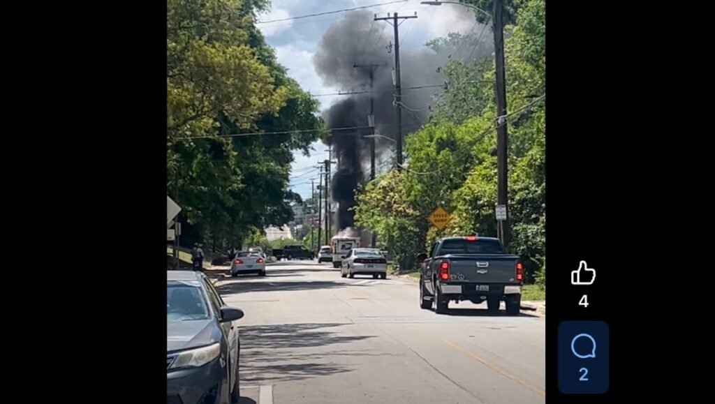A street scene with several vehicles parked along the side and a few more driving. In the background, thick black smoke rises from a location beyond some trees, indicating a possible fire or incident.