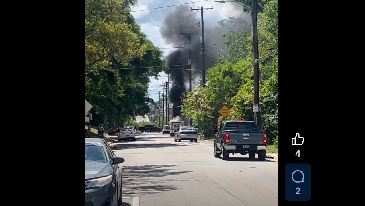 A street scene with several vehicles parked along the side and a few more driving. In the background, thick black smoke rises from a location beyond some trees, indicating a possible fire or incident.