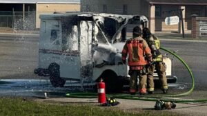 Firefighters extinguishing a burning postal delivery truck. The vehicle has heavy fire damage, with smoke and water spraying from the rear. Nearby, a fire extinguisher is positioned on the ground, and a fire hose is being used in the operation.