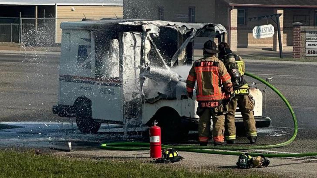 Firefighters extinguishing a burning postal delivery truck. The vehicle has heavy fire damage, with smoke and water spraying from the rear. Nearby, a fire extinguisher is positioned on the ground, and a fire hose is being used in the operation.