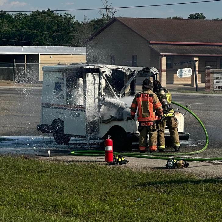 Firefighters extinguishing a burnt delivery vehicle with visible smoke and water spraying from hoses. The charred vehicle is parked on a roadside near a commercial building, with fire fighting equipment and personnel in uniform nearby.