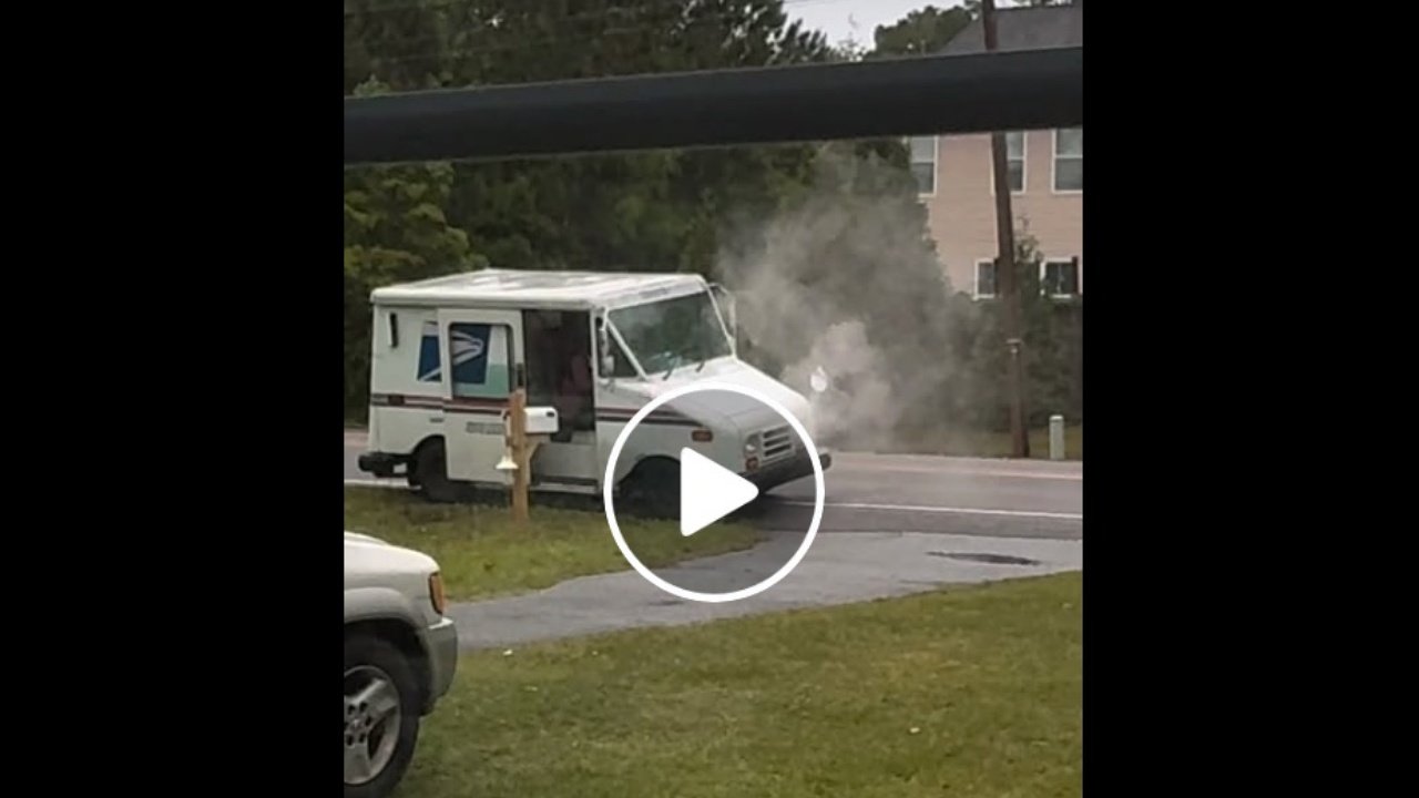 A U.S. Postal Service delivery truck is seen parked on a road with smoke billowing from the front hood. The background features a grassy area and scattered trees, while a vehicle is partially visible in the foreground.