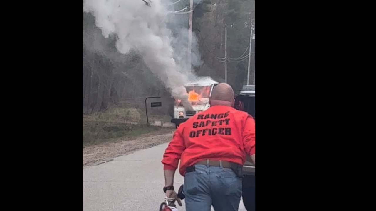A person wearing a red jacket labeled 'RANGE SAFETY OFFICER' stands on a road, facing a vehicle engulfed in smoke and flames. Power lines are visible in the background, along with trees lining the roadside.