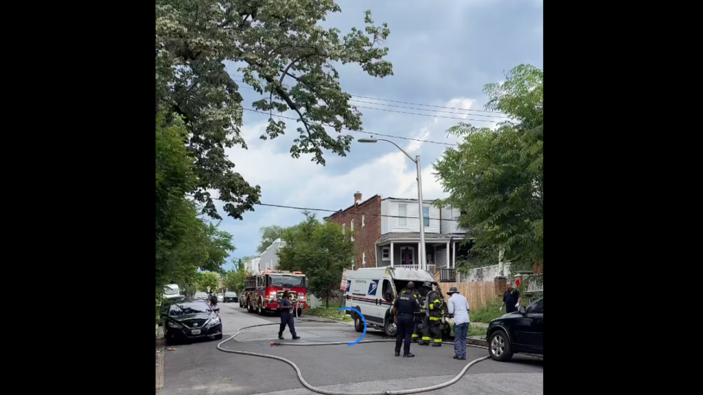 Firefighters responding to an incident on a residential street. Several emergency vehicles are parked nearby, including a fire truck and an ambulance. Firefighters are seen directing a hose while onlookers watch from the sidewalk.