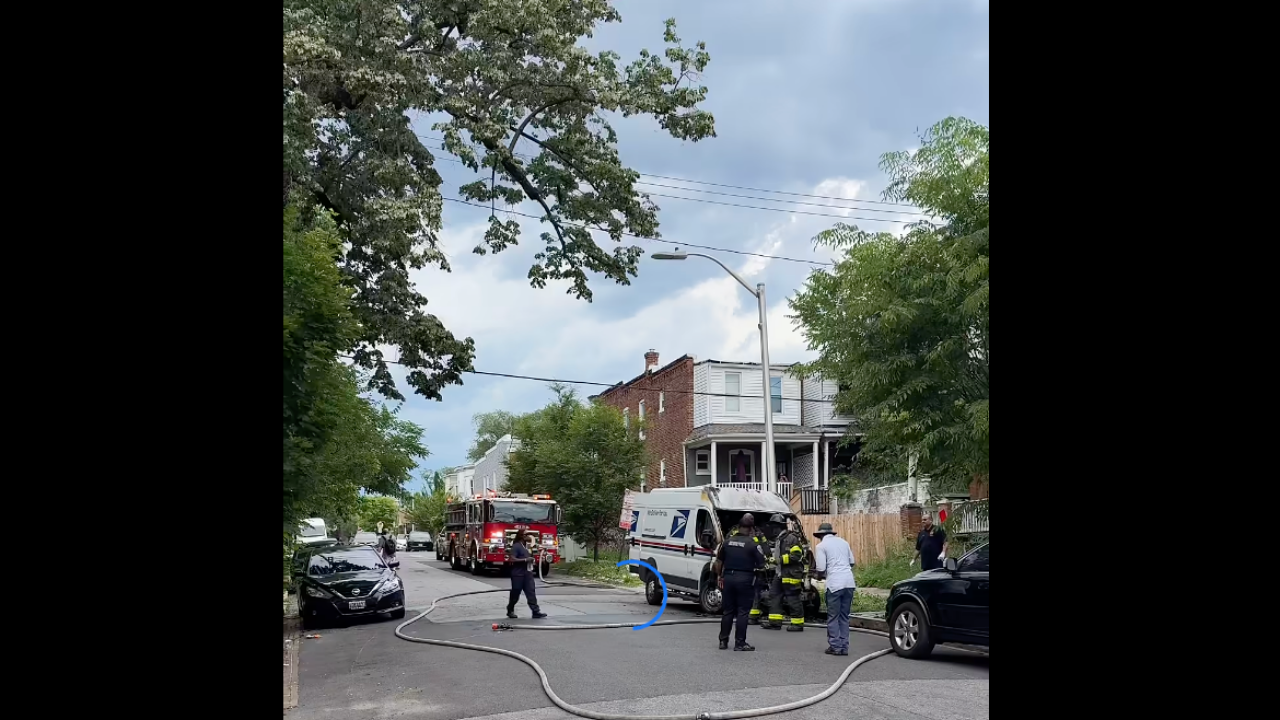 Firefighters responding to an incident on a residential street. Several emergency vehicles are parked nearby, including a fire truck and an ambulance. Firefighters are seen directing a hose while onlookers watch from the sidewalk.