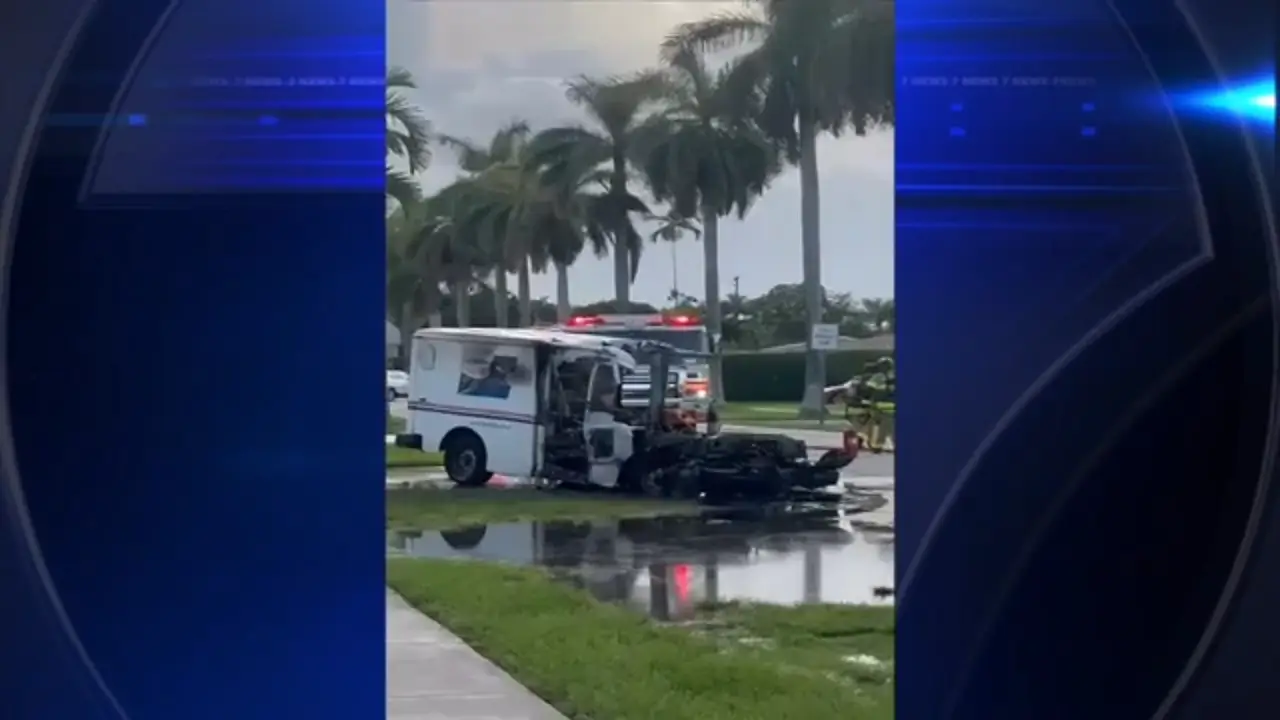 A white postal service truck is damaged and partially overturned on the side of the road, with visible water pooling around it. Palm trees and a residential area are in the background, while emergency lights can be seen on a vehicle nearby.