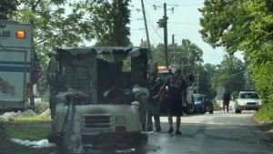 A fire-damaged vehicle is parked on the side of a road, with firefighters inspecting the scene. An ambulance is visible in the background, along with other vehicles and people walking nearby, surrounded by trees and a clear sky.