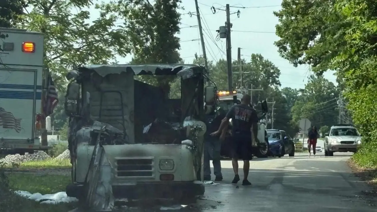A fire-damaged vehicle is parked on the side of a road, with firefighters inspecting the scene. An ambulance is visible in the background, along with other vehicles and people walking nearby, surrounded by trees and a clear sky.