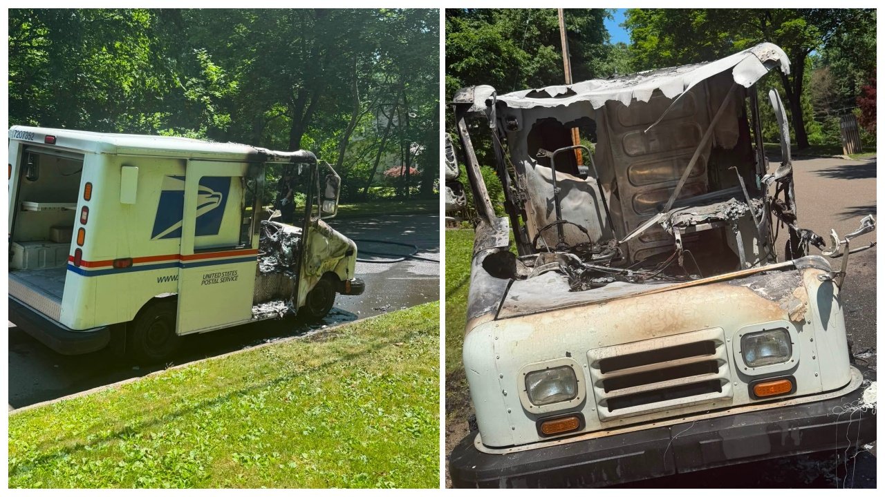 A damaged United States Postal Service truck shows significant fire damage. The rear of the vehicle is intact, while the front is heavily charred with a melted hood and burnt interior. The setting includes lush green grass and a street lined with trees.