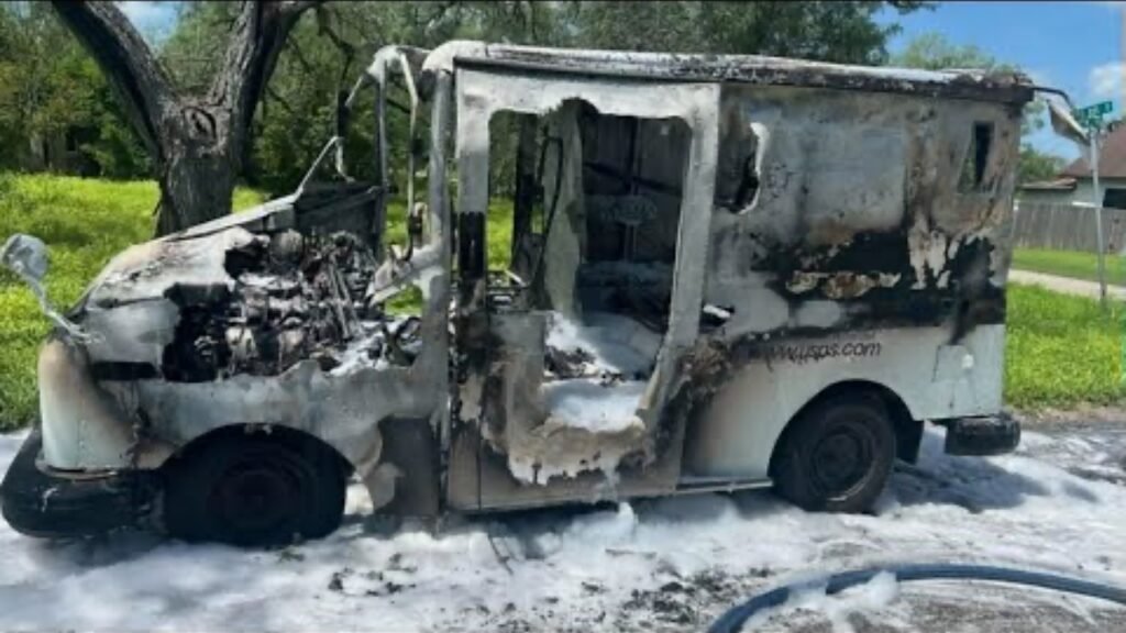 A burnt delivery truck with charred metal and exposed components is parked on the street. Firefighting foam covers the ground around the vehicle, and green grass and trees are visible in the background.