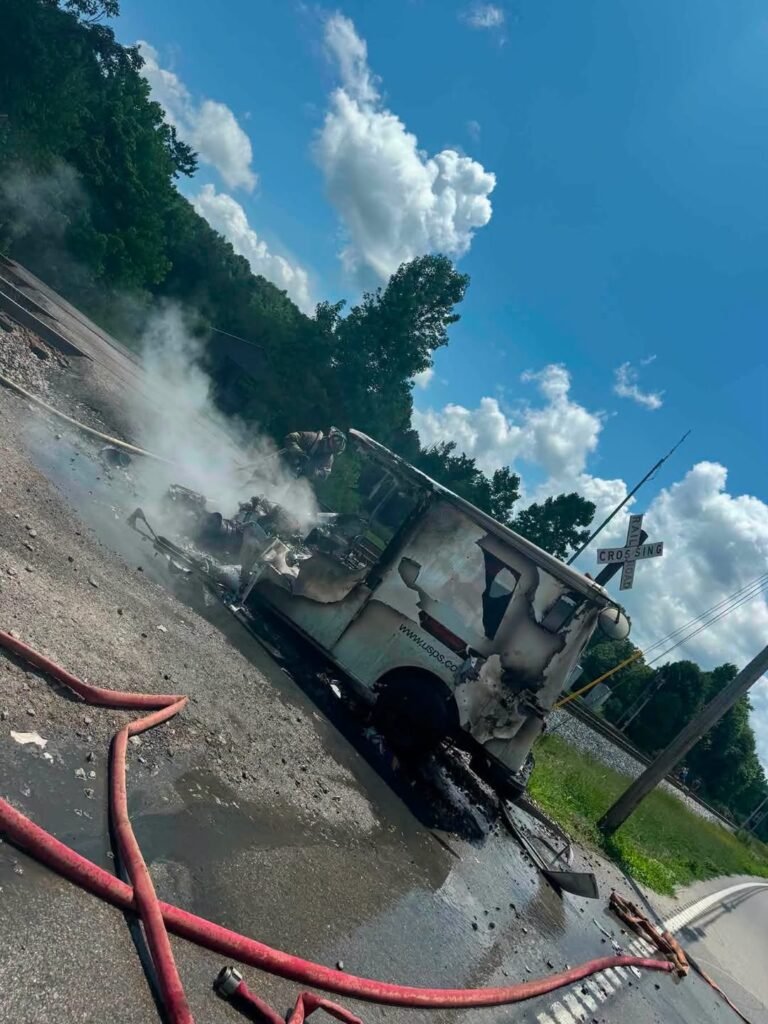 A burned and damaged food truck is parked near a railroad crossing, emitting smoke. Fire hoses lie on the ground in the foreground, and the background features green trees and a partly cloudy blue sky.