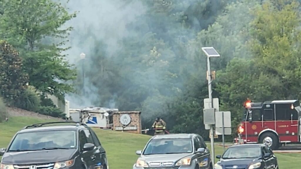 A fire truck is parked near a smoke-emitting structure on a grassy area. Several cars are visible in the foreground, while emergency personnel are attending to the scene amidst greenery in the background.