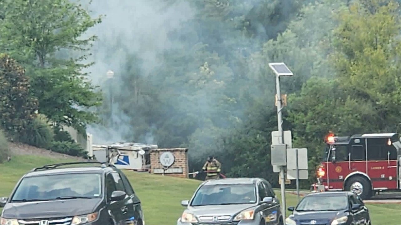 A fire truck is parked near a smoke-emitting structure on a grassy area. Several cars are visible in the foreground, while emergency personnel are attending to the scene amidst greenery in the background.
