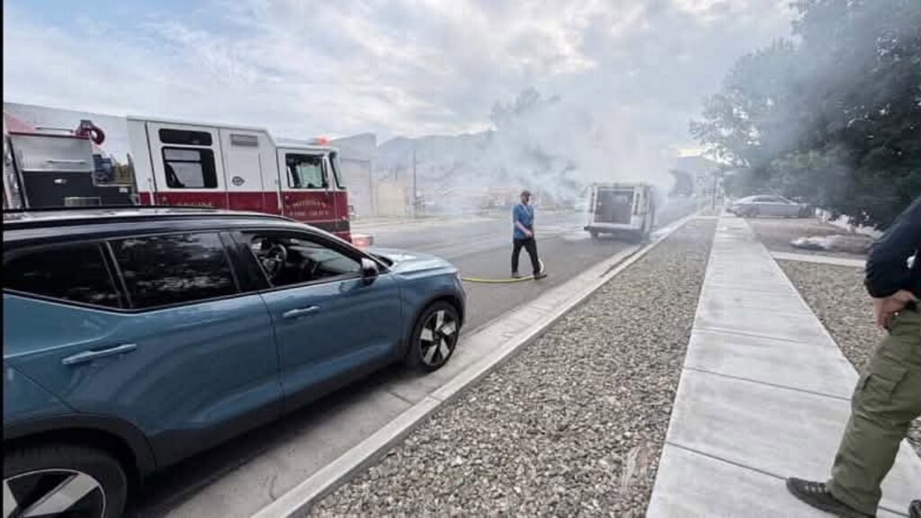 A blue SUV is parked near a fire truck responding to an emergency. Smoke fills the air as a man in a blue shirt stands near a van, while another person approaches from the sidewalk on a street lined with trees.