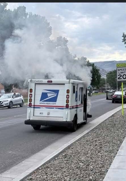 A United States Postal Service delivery truck is parked on a road, emitting a large cloud of smoke from its rear. Nearby vehicles are visible, and a school zone sign indicates a speed limit of 20 mph.