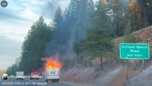 A USPS delivery truck on the side of a road is engulfed in flames, producing black smoke. Nearby, a green directional sign indicates "Allison Ranch Road, Next Right," with trees in the background and a clear sky above.