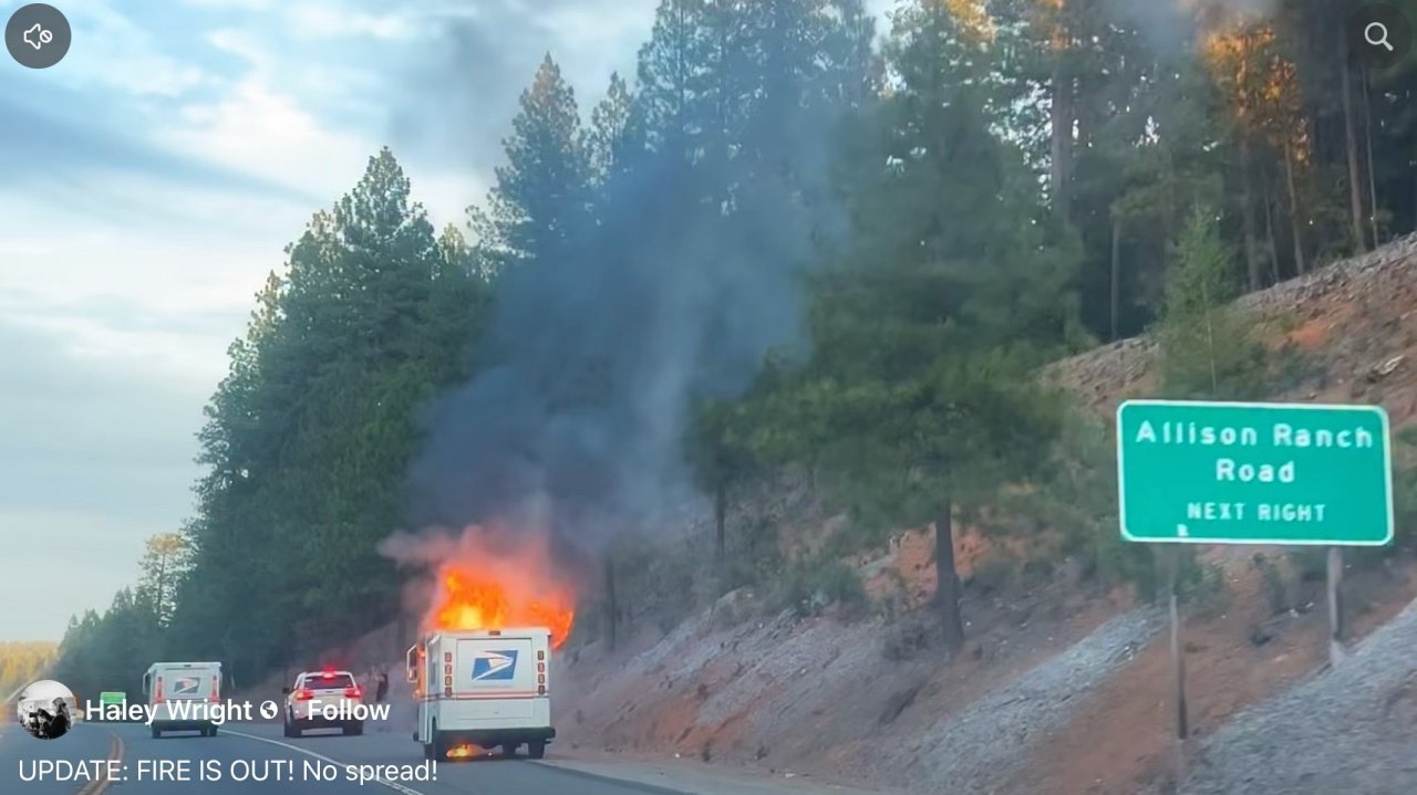 A USPS delivery truck on the side of a road is engulfed in flames, producing black smoke. Nearby, a green directional sign indicates "Allison Ranch Road, Next Right," with trees in the background and a clear sky above.