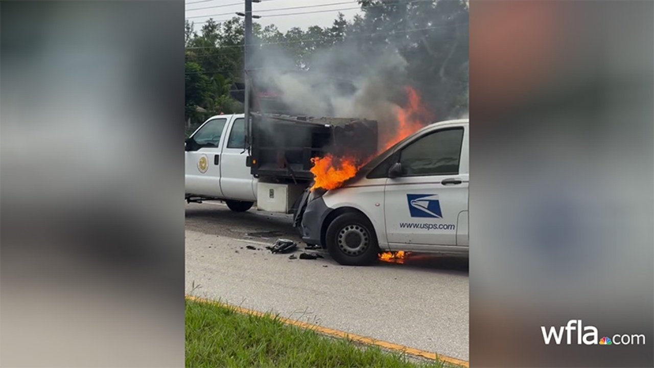 A postal van is on fire in a two-lane road, with flames and smoke billowing from the front. A white pickup truck is parked nearby, and green grass lines the side of the road. Utility poles and trees are visible in the background.