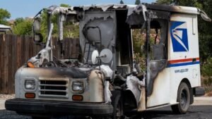 A burned-out USPS mail truck with a badly damaged front and roof sits on a street beside a wooden fence and trees.