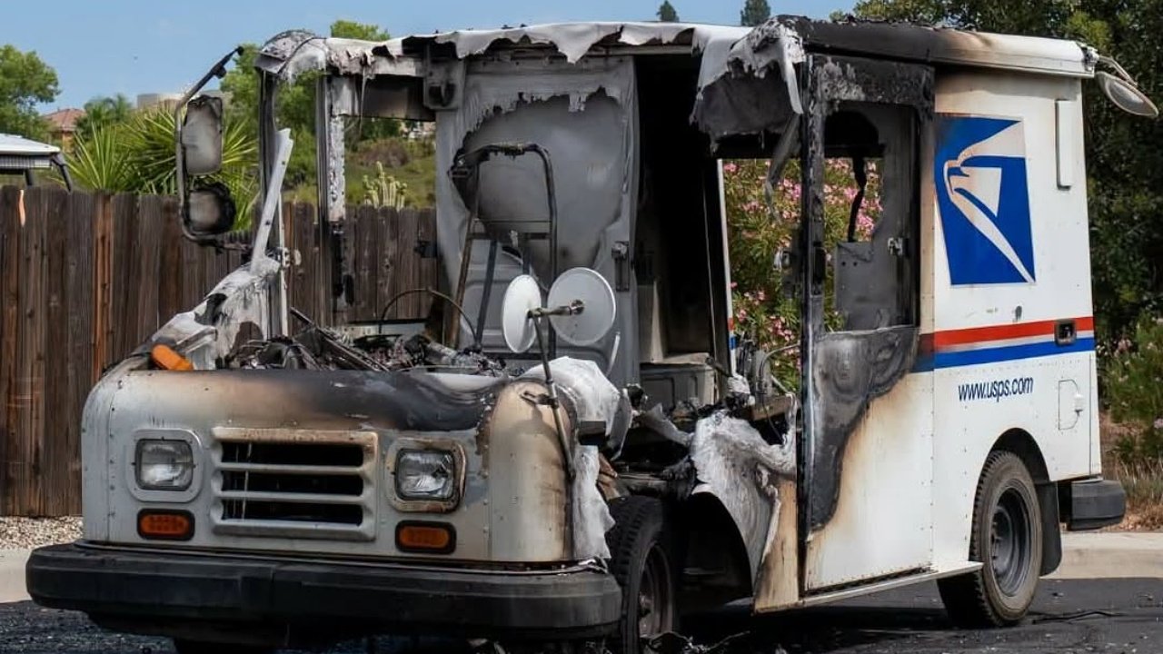 A burned-out USPS mail truck with a badly damaged front and roof sits on a street beside a wooden fence and trees.