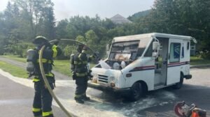 Three firefighters in protective gear spray water into the front of a white delivery van parked on a wet road, with trees and a house in the background.