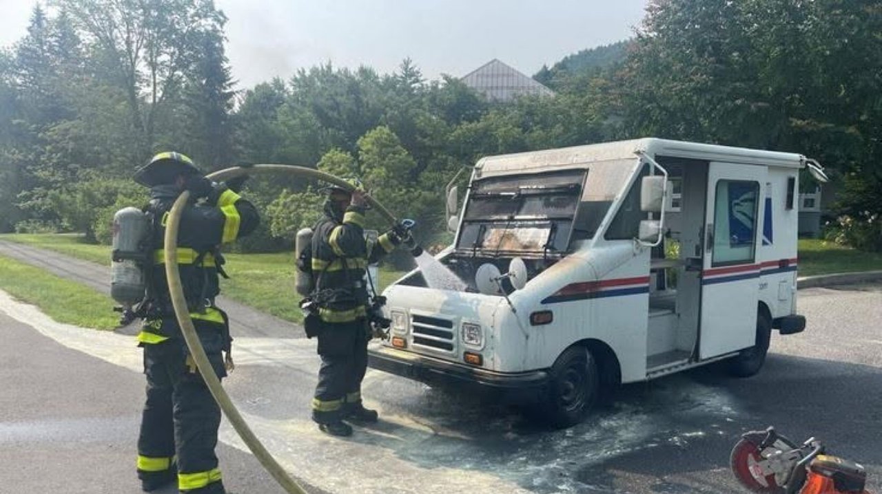 Three firefighters in protective gear spray water into the front of a white delivery van parked on a wet road, with trees and a house in the background.