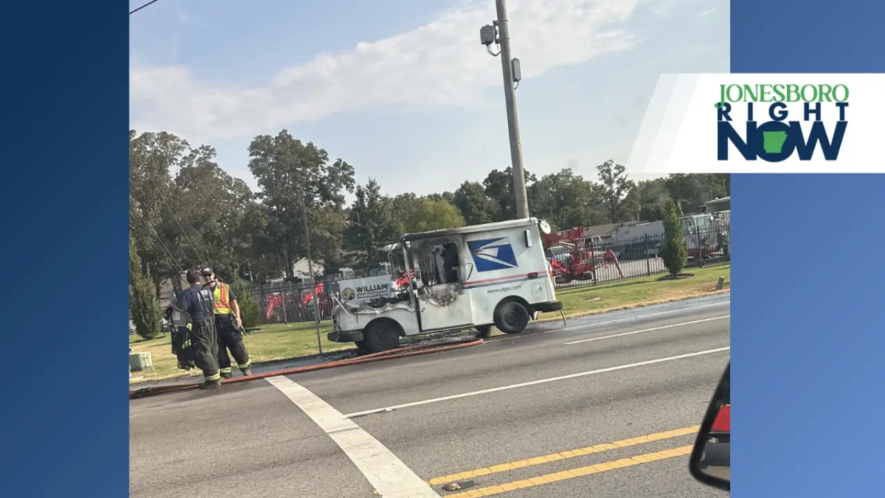 A white USPS mail truck sits on the roadside near a utility pole and several firefighters, with trees and emergency vehicles in the background.