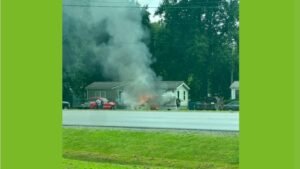 A house with a fire and thick gray smoke rising from the roof, with a red pickup truck and trees in the background.